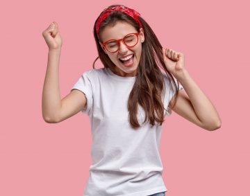 Triumphing joyful woman clenches fists, rejoices positive news, exclaims gladfully, models over pink background, achieves victory and success, isolated over pink background. Overemotive teenage girl