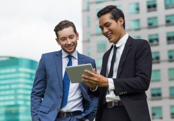 Closeup of two smiling young business men using tablet computer and standing with office buildings in background