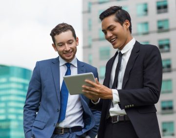 Closeup of two smiling young business men using tablet computer and standing with office buildings in background