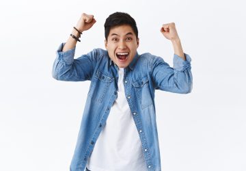 Waist-up portrait of happy, excited young smiling man shouting hooray or yes, raising hands up, fist pump in joy, rejoicing over win, achieve goal, celebrate victory or success, white background.