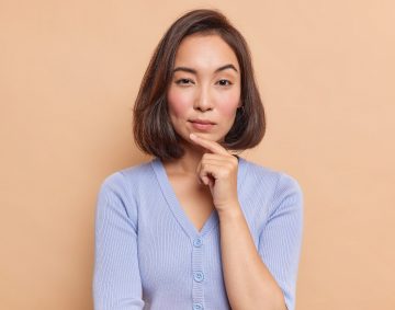 Portrait of serious dark haired Asian woman keeps finger on chin looks mysteriously at camera considers something dressed in blue jumper isolated over brown background. Let me think about it